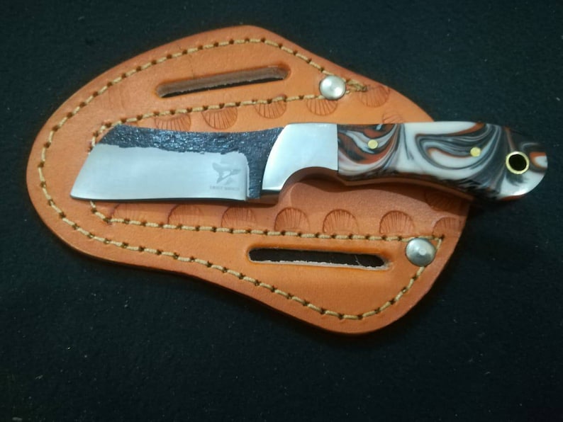 May include: A small, stainless steel knife with a marbled white, black, and brown handle, resting in a tan leather sheath. The blade has a dark, textured edge and the text "Eagle Knife".