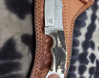 May include: A hunting knife with a stainless steel blade and a textured handle made of antler material. The knife is in a brown leather sheath with decorative stitching and a ring at the base of the handle.