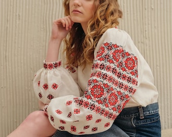 May include: A woman wearing a white blouse with red and black embroidery and a blue denim skirt. She is sitting with her hand on her chin and looking to the side.