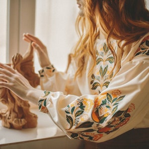 Puede incluir: Una mujer con una blusa blanca con bordados florales. La blusa tiene mangas largas y un escote redondo. El bordado es en tonos de verde, amarillo y naranja.