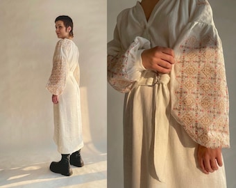 Pode incluir: Um vestido branco de linho com mangas bordadas. As mangas apresentam um padrão geométrico em tons de marrom, vermelho e branco. O vestido tem uma cintura ajustada e uma saia fluida.