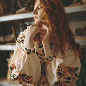 Puede incluir: Una mujer con el pelo largo y rojo lleva una blusa blanca con estampado floral y una falda marr&oacute;n. La blusa tiene mangas largas con un pu&ntilde;o fruncido. La mujer est&aacute; de pie frente a un estante con varios objetos en &eacute;l.