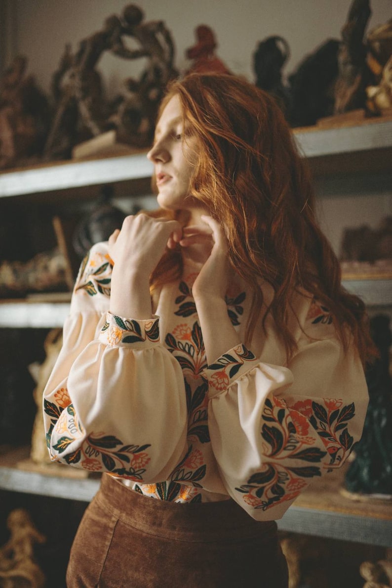 Puede incluir: Una mujer con el pelo largo y rojo lleva una blusa blanca con estampado floral y una falda marr&oacute;n. La blusa tiene mangas largas con un pu&ntilde;o fruncido. La mujer est&aacute; de pie frente a un estante con varios objetos en &eacute;l.