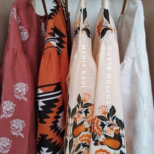 Puede incluir: Cuatro blusas bordadas colgadas de perchas de madera. Las blusas son de varios colores, incluyendo rojo, naranja y blanco. Los dise&ntilde;os de bordado incluyen flores, p&aacute;jaros y patrones geom&eacute;tricos. Las etiquetas en las blusas indican la composici&oacute;n de la tela: "100% lino" y "mezcla de algod&oacute;n".