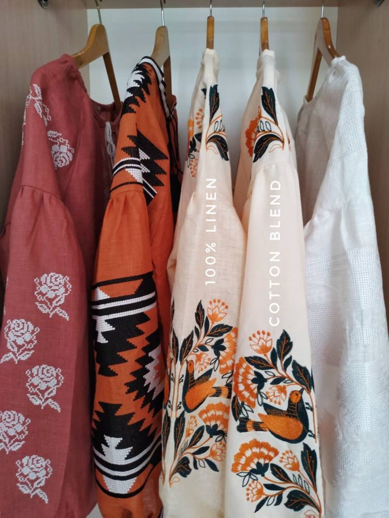 Puede incluir: Cuatro blusas bordadas colgadas de perchas de madera. Las blusas son de varios colores, incluyendo rojo, naranja y blanco. Los dise&ntilde;os de bordado incluyen flores, p&aacute;jaros y patrones geom&eacute;tricos. Las etiquetas en las blusas indican la composici&oacute;n de la tela: "100% lino" y "mezcla de algod&oacute;n".