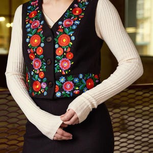 May include: A black vest with colorful floral embroidery. The vest has a V-neck and a button closure.