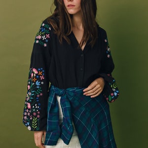 Puede incluir: Una camisa negra de botones con detalles florales bordados en las mangas. La camisa se lleva con una minifalda blanca y una camisa de cuadros azules y verdes atada a la cintura.