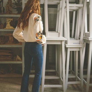 Puede incluir: Una mujer con una blusa blanca con bordados florales y pantalones vaqueros azules se encuentra frente a una pila de sillas blancas.