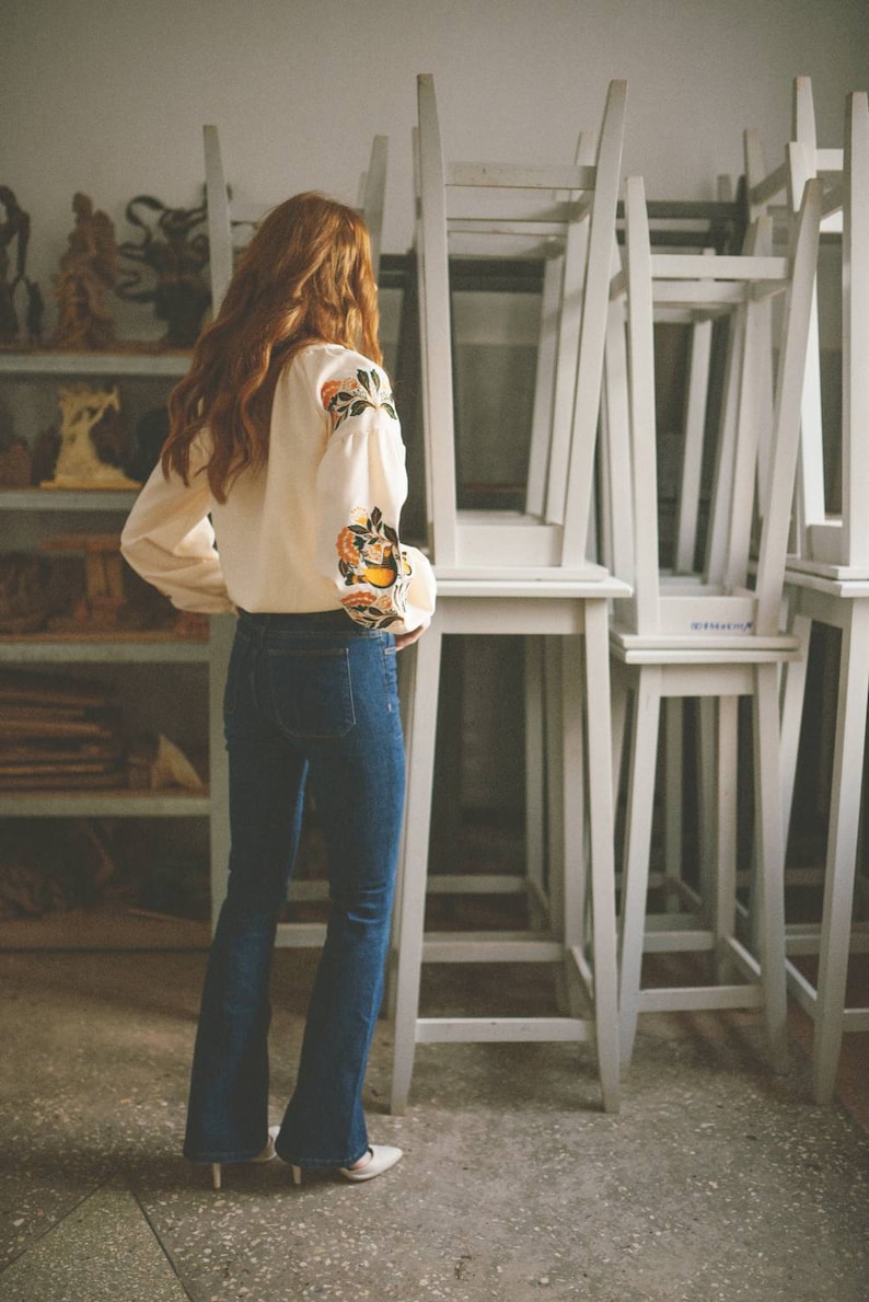Puede incluir: Una mujer con una blusa blanca con bordados florales y pantalones vaqueros azules se encuentra frente a una pila de sillas blancas.