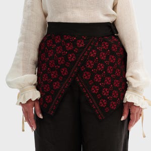 Peut inclure: Une jupe portefeuille noire avec broderie florale rouge. La jupe a une ceinture noire et est portée avec un pantalon noir et un chemisier crème à manches bouffantes.