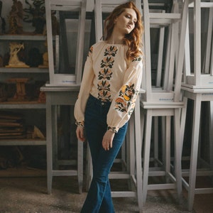 Puede incluir: Una mujer con una blusa blanca bordada con un patr&oacute;n floral y pantalones vaqueros azules. Est&aacute; de pie frente a una pila de sillas blancas.
