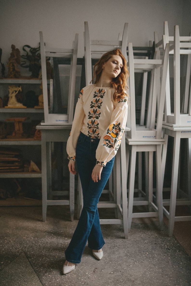 Puede incluir: Una mujer con una blusa blanca bordada con un patr&oacute;n floral y pantalones vaqueros azules. Est&aacute; de pie frente a una pila de sillas blancas.