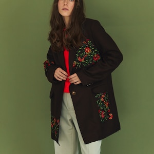 May include: A woman wearing a long, dark brown coat with floral embroidery. The coat has a red button closure and a pocket on the left side. The woman is wearing white pants.