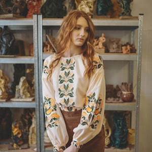 Puede incluir: Una mujer con el pelo largo y rojo lleva una blusa blanca con bordados florales verdes y amarillos y una falda de ante marr&oacute;n. Est&aacute; de pie frente a una estanter&iacute;a con varias esculturas.