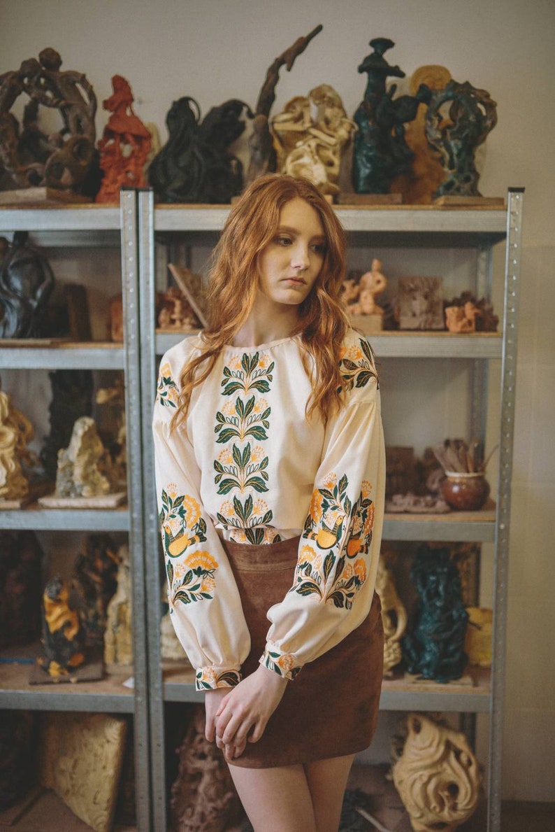 Puede incluir: Una mujer con el pelo largo y rojo lleva una blusa blanca con bordados florales verdes y amarillos y una falda de ante marr&oacute;n. Est&aacute; de pie frente a una estanter&iacute;a con varias esculturas.