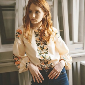 Puede incluir: Una mujer con el pelo largo y rojo lleva una blusa blanca con bordados florales y pantalones vaqueros azules. La blusa tiene un cuello redondo y mangas largas. El bordado es en tonos naranja, verde y amarillo.