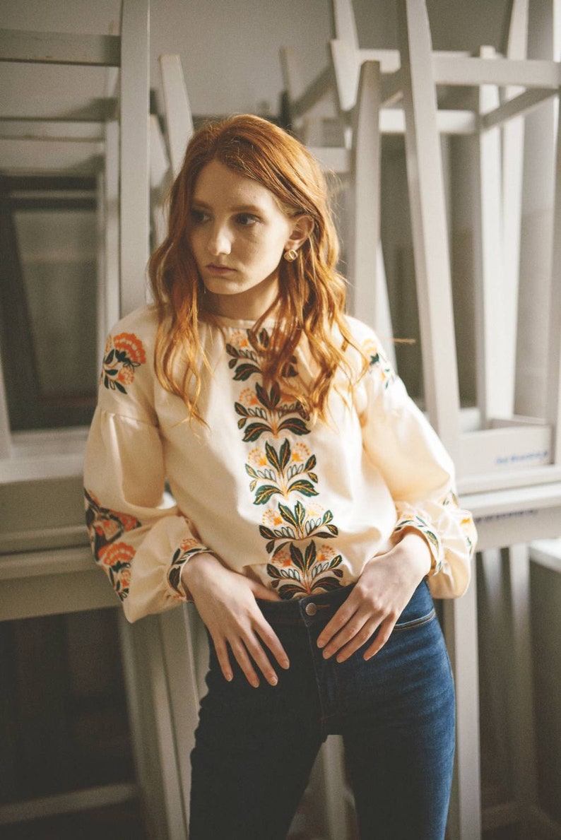 Puede incluir: Una mujer con el pelo largo y rojo lleva una blusa blanca con bordados florales y pantalones vaqueros azules. La blusa tiene un cuello redondo y mangas largas. El bordado es en tonos naranja, verde y amarillo.