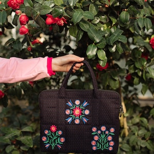 May include: A black laptop bag with floral embroidery. The bag has a zipper closure and two handles. The embroidery is in shades of pink, blue, and green.