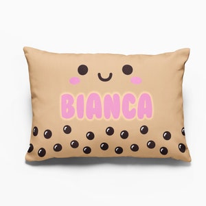 May include: A tan pillow with a cute cartoon face and the name "BIANCA" in pink letters. The pillow has a pattern of black boba tea pearls.