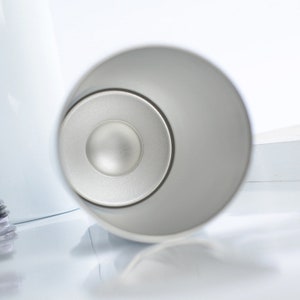 May include: A close-up shot of a silver-colored object with a circular design. The object has a smooth, metallic finish and a central, slightly raised disc. The background is a bright white, creating a high-contrast image.
