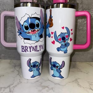 May include: Two white travel mugs with pink and purple handles. The mugs feature a blue cartoon character with big eyes and a wide smile. One mug has the name "BRYNLYN" on it.