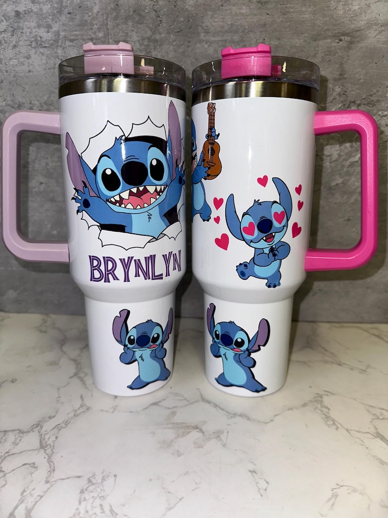 May include: Two white travel mugs with pink and purple handles. The mugs feature a blue cartoon character with big eyes and a wide smile. One mug has the name "BRYNLYN" on it.