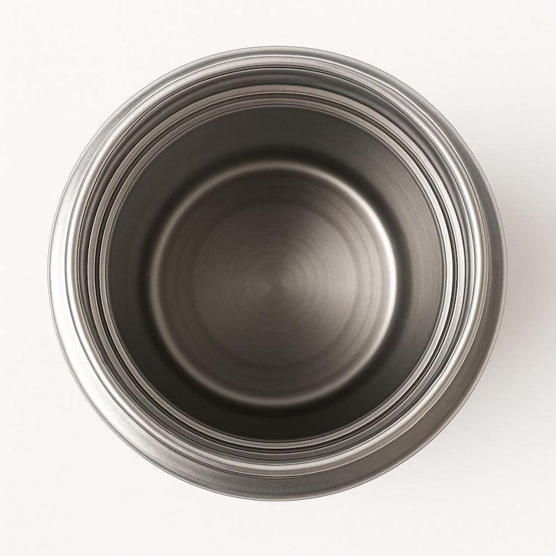 May include: A stainless steel pot, viewed from above, with a circular design. The pot has a shiny, metallic finish and a simple, modern aesthetic. The interior is a darker shade of gray, with concentric circles.