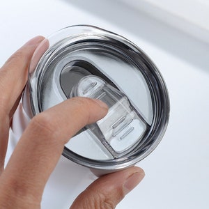 May include: Close-up of a white tumbler with a silver rim and a clear plastic lid. A finger is pressing down on the lid's opening mechanism. The tumbler appears to be made of stainless steel.