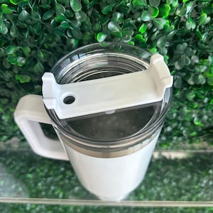 May include: White travel mug with a lid and a handle. The mug has a stainless steel band around the middle.