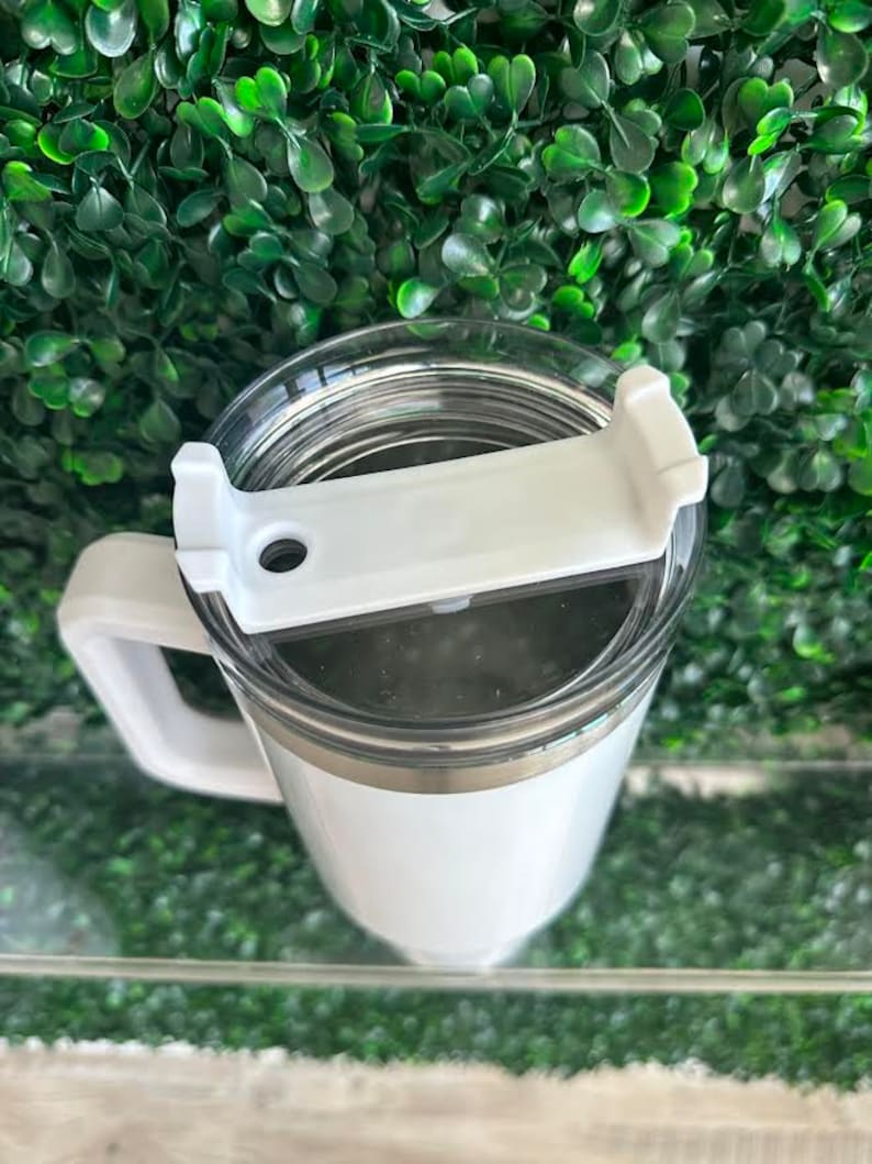May include: White travel mug with a lid and a handle. The mug has a stainless steel band around the middle.