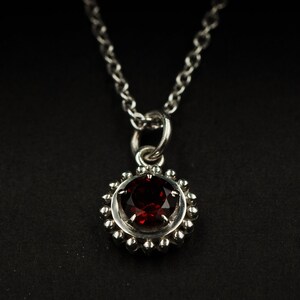 May include: A silver necklace with a red gemstone pendant. The pendant is surrounded by a circle of small silver beads.