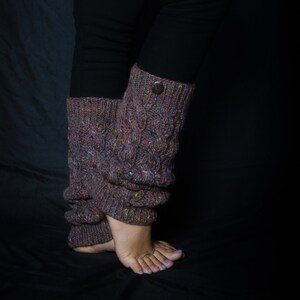May include: Knit leg warmers in a textured cable pattern, featuring a button detail. The leg warmers are a mix of brown, purple, and colorful flecks. The person is wearing black leggings. The leg warmers are designed to be worn around the ankles and lower legs.