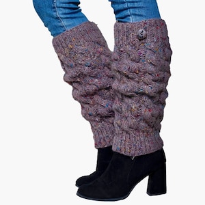 May include: A pair of mauve-colored cable knit leg warmers with a button detail, worn over black boots and under blue jeans. The leg warmers have flecks of colorful yarn throughout.