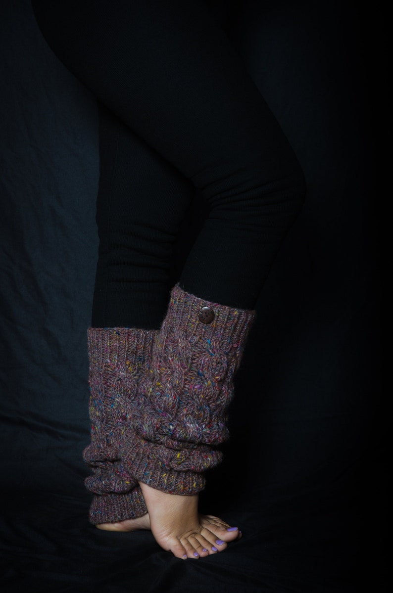 May include: Knit leg warmers in a textured cable pattern, featuring a button detail. The leg warmers are a mix of brown, purple, and colorful flecks. The person is wearing black leggings. The leg warmers are designed to be worn around the ankles and lower legs.