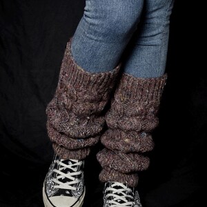 May include: A person wearing blue jeans and brown cable-knit leg warmers over black and white patterned sneakers. The leg warmers are a textured, chunky knit with flecks of color.