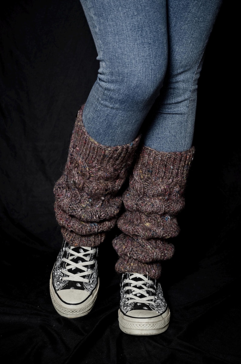 May include: A person wearing blue jeans and brown cable-knit leg warmers over black and white patterned sneakers. The leg warmers are a textured, chunky knit with flecks of color.