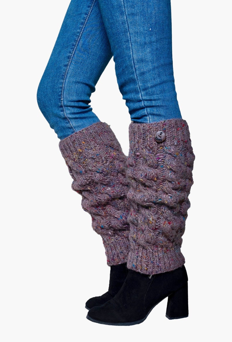 May include: A pair of mauve-colored cable knit leg warmers with a button detail, worn over black boots and under blue jeans. The leg warmers have flecks of colorful yarn throughout.