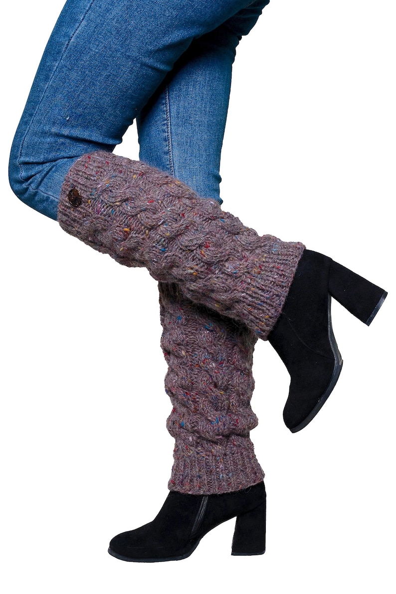 May include: A pair of purple leg warmers with a cable knit design and a button detail. The leg warmers are worn with blue jeans and black heeled boots. The knitwear has flecks of color throughout.