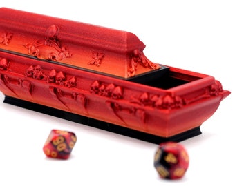 Könnte beinhalten: Eine rote, sargförmige Würfelbox mit Totenkopf- und gekreuzten Knochen-Details. Die Box ist geöffnet und zeigt einen schwarzen Innenraum. Mehrere vielseitige Würfel mit roter, schwarzer und goldener Farbe sind um die Box herum verstreut.