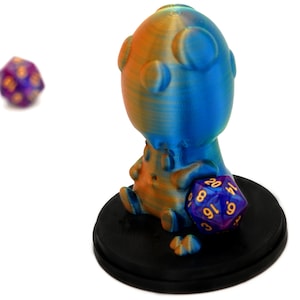 Pode incluir: Um porta-dados impresso em 3D azul e laranja em forma de criatura com uma cabeça grande e um corpo pequeno. O porta-dados está sobre uma base preta e tem um dado roxo e dourado na mão.