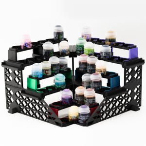 May include: A black paint bottle organiser with multiple tiers, holding various paint bottles. The organiser has green, red, and purple accents. The bottles contain paints in colours such as pink, green, and brown.