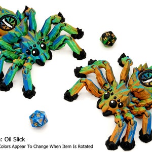 May include: Two 3D printed spider figurines with iridescent paint. The spiders have eight legs, a segmented body, and a large eye on their back. The spiders are painted in shades of green, blue, and gold. The text "Quantum: Oil Slick" and "*Quantum Colors Appear To Change When Item Is Rotated" is visible below the spiders.