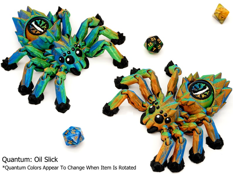 May include: Two 3D printed spider figurines with iridescent paint. The spiders have eight legs, a segmented body, and a large eye on their back. The spiders are painted in shades of green, blue, and gold. The text "Quantum: Oil Slick" and "*Quantum Colors Appear To Change When Item Is Rotated" is visible below the spiders.