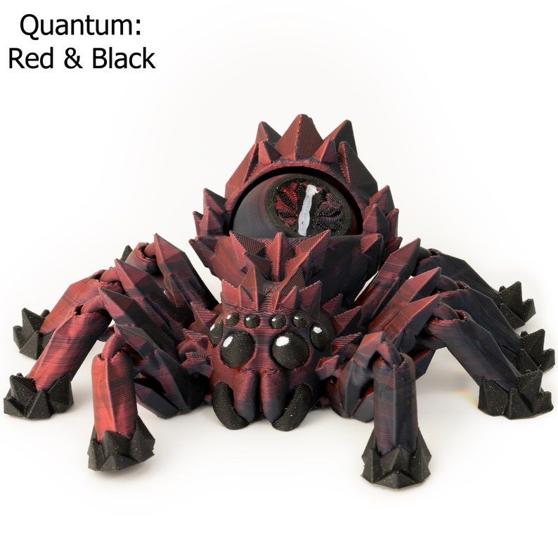 May include: A red and black 3D printed spider with a large, round eye in the center of its head. The spider has eight legs and a spiky body. The text "Quantum: Red & Black" is visible in the top left corner of the image.