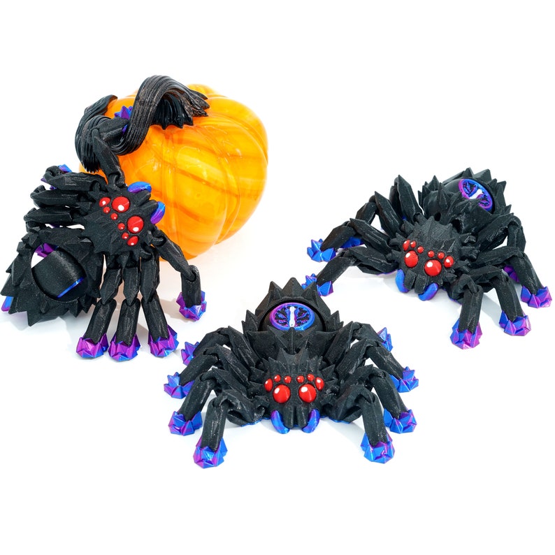 May include: Three black spiders with red eyes and purple accents. The spiders have eight legs and are posed on a white surface. A small orange pumpkin is also in the image.