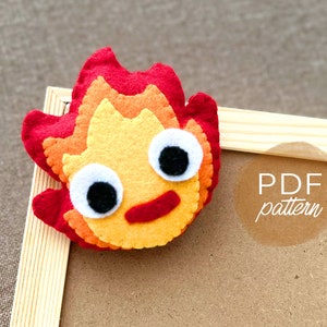 May include: A felt plush toy shaped like a fire spirit with a red, orange, and yellow flame design. The toy has large white eyes with black pupils and a red mouth. The toy is sitting on a wooden frame with the text "PDF pattern" written on it.
