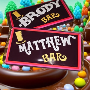 May include: Two chocolate bar wrappers with personalised names, "Brody Bar" and "Matthew Bar", on a chocolate cake with colourful candies.