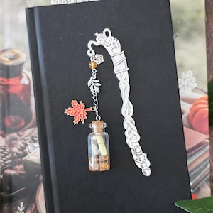 Puede incluir: Un marcapáginas de metal plateado con un intrincado diseño trenzado y una cadena de dijes colgantes. La cadena incluye un pequeño dije de hoja de arce naranja y una botella de vidrio en miniatura llena de diminutos pergaminos y cuentas ámbar.
