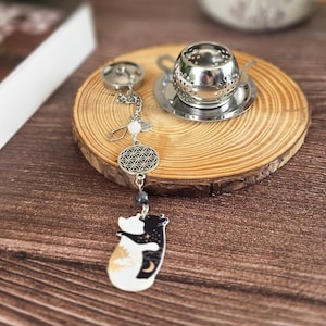 Peut inclure: Un infuseur à thé en acier inoxydable avec une chaînette ornée d'une breloque fleur de vie argentée, une perle blanche et un pendentif plat représentant un chat blanc et un chat noir s'enlaçant, avec des motifs de soleil et de lune sur leurs corps.