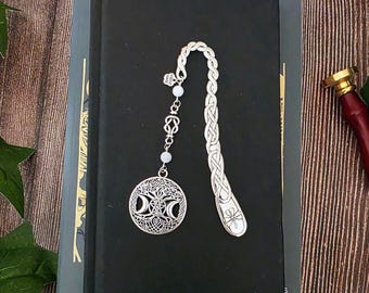 Op de afbeelding: Een zilverkleurige metalen boekenlegger met een gevlochten handvat en een bungelende ronde bedel. De bedel toont een centraal pentagram omringd door maansikkels en ingewikkeld knoopwerk. Twee kleine witte kralen verbinden de bedel met het handvat.